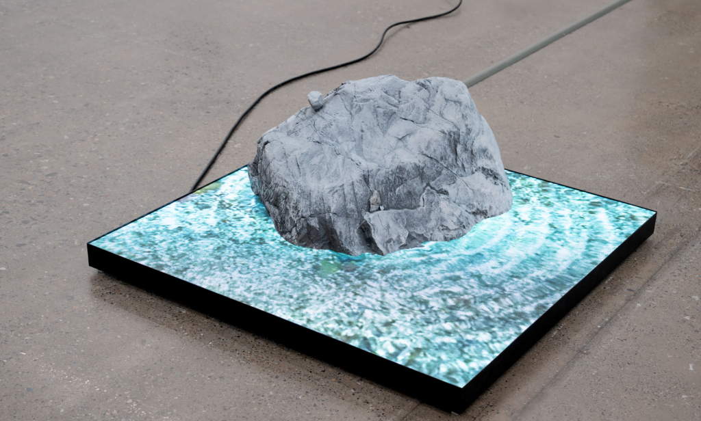 an artwork that looks like a rock sitting in water, the water is made of a digital screen playing water footage